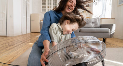 Playful single mother bonding with son excited playing together at home with big fan blowing cool wind in living room. Mum and child enjoy time together at conditioning ventilator during summer heat